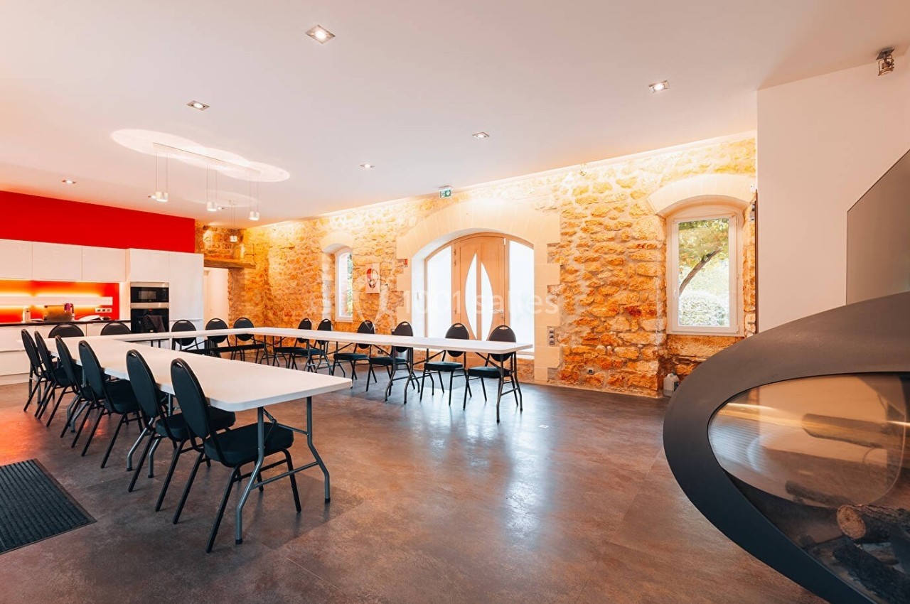 Salle lumineuse avec murs en pierre, tables blanches alignées, chaises noires et cuisine moderne en arrière-plan.