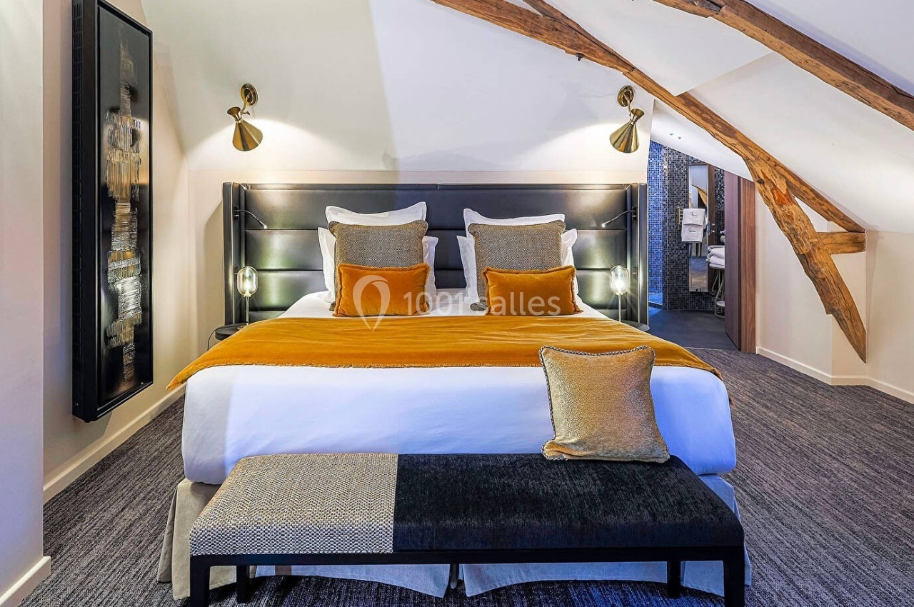 Chambre mansardée avec lit double, coussins jaunes, banc au pied du lit et éclairage moderne sous poutres apparentes.