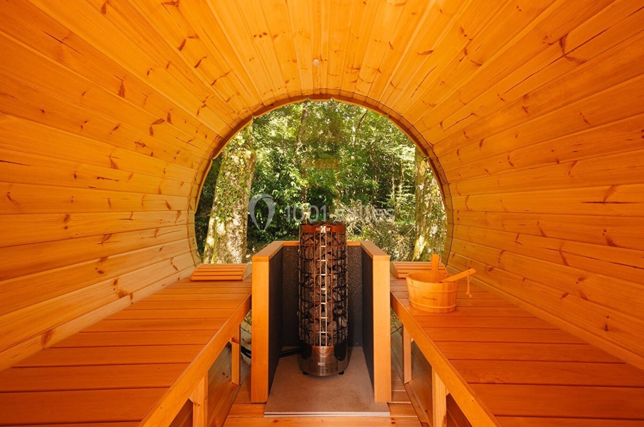 Intérieur d'un sauna en bois en forme de tonneau, avec un poêle central et une vue sur un environnement boisé.