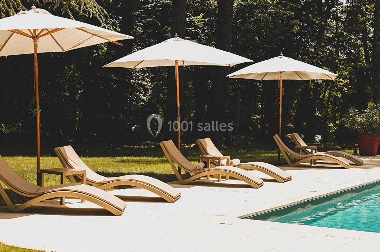 Chaises longues et parasols alignés au bord d'une piscine, entourés de verdure et d'arbres.