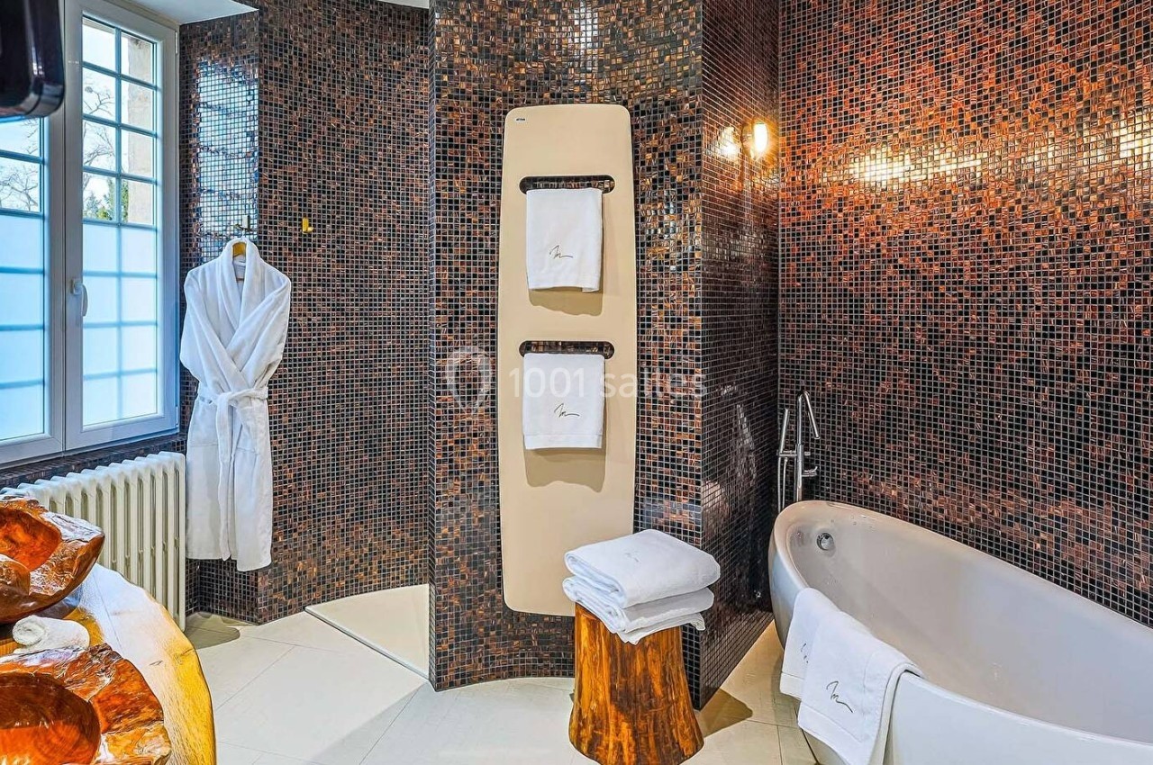 Salle de bain avec mosaïque sombre, baignoire, porte-serviettes, lavabo en bois sculpté et peignoir suspendu.