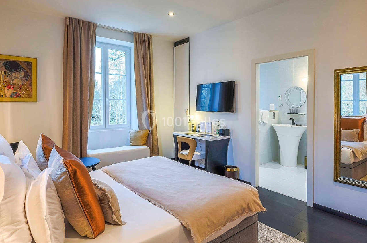 Chambre lumineuse avec lit double, bureau, télévision, grande fenêtre et salle de bain attenante visible.