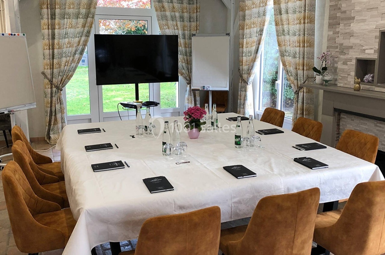 Salle de réunion lumineuse avec table rectangulaire dressée, chaises en velours, écran, paperboards et vue sur jardin.