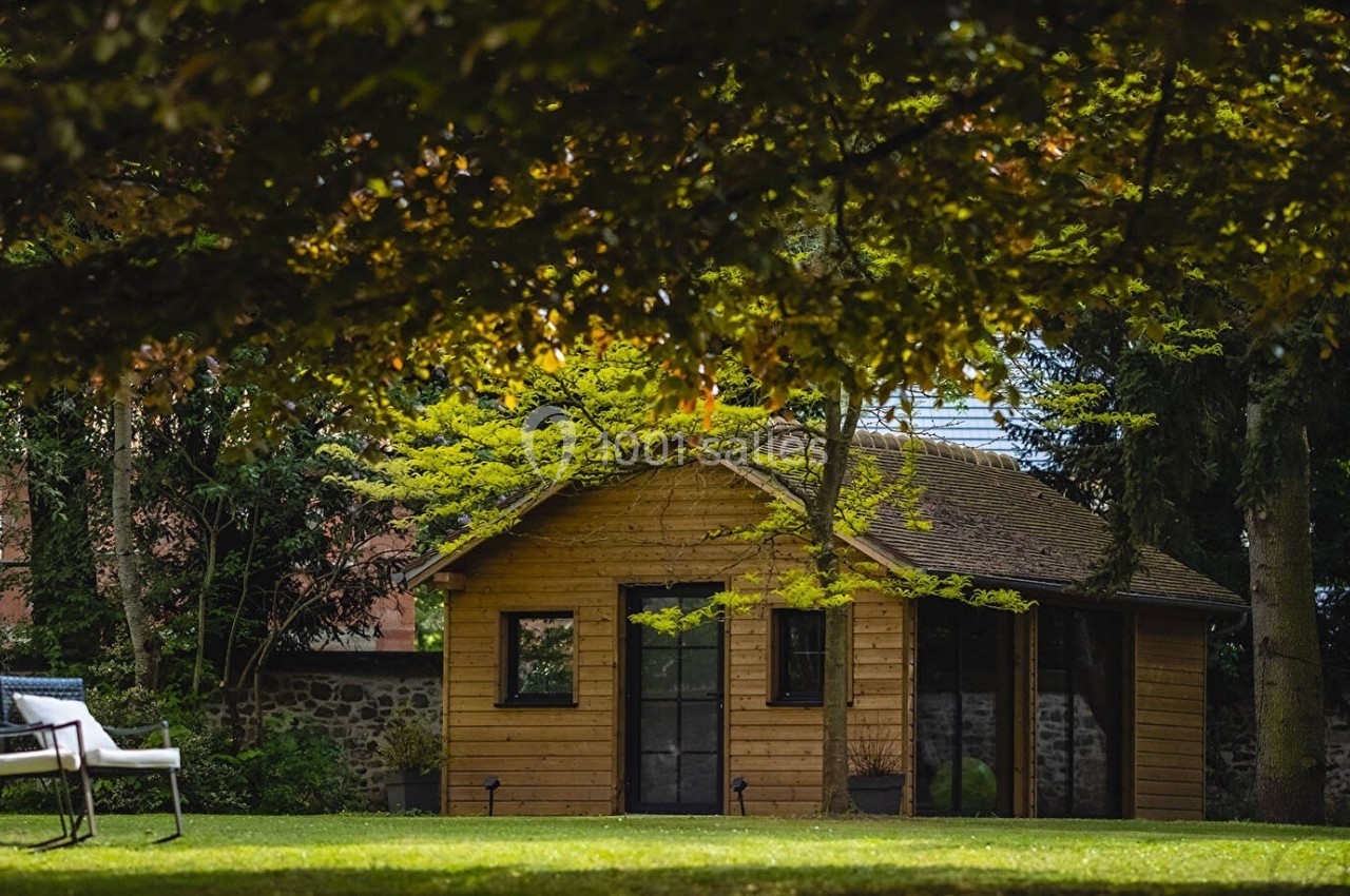Abri de jardin en bois entouré de verdure, avec des arbres et une chaise blanche sur la pelouse au premier plan.