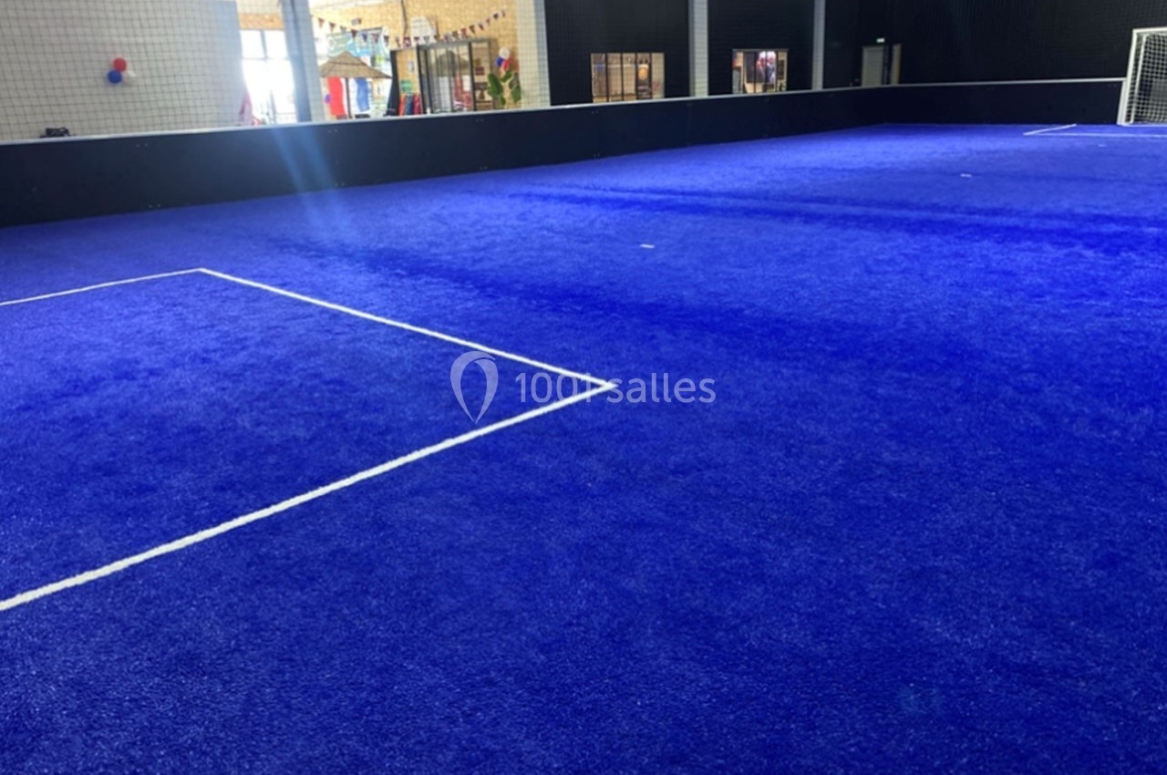 Terrain de sport intérieur avec revêtement bleu, lignes blanches marquant les limites et un but visible en arrière-plan.