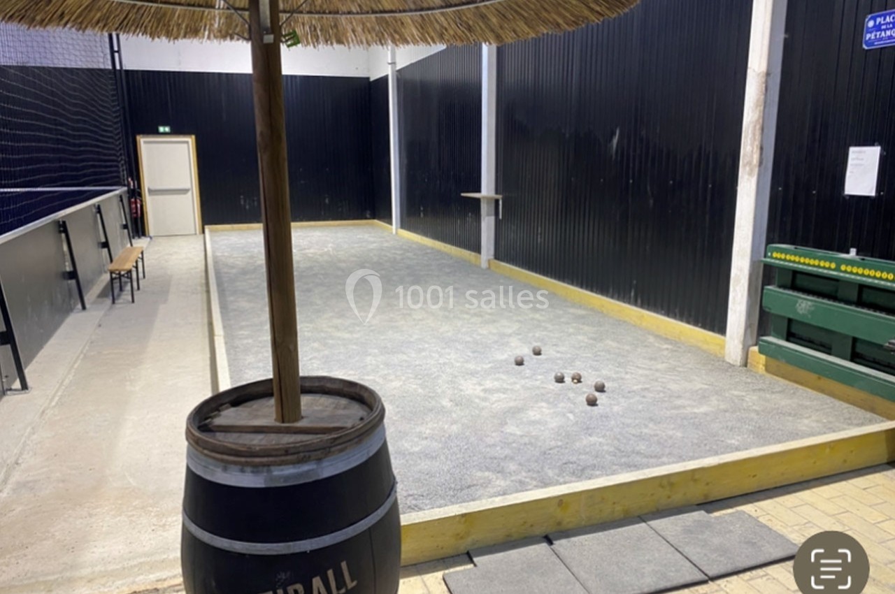 Terrain de pétanque intérieur avec sol en gravier, quelques boules disposées et un tonneau en premier plan.
