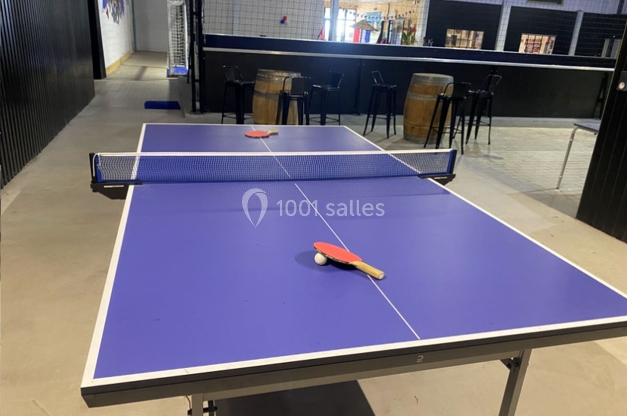Table de tennis de table bleue avec deux raquettes rouges posées, dans un espace intérieur avec des tables hautes en arrière…