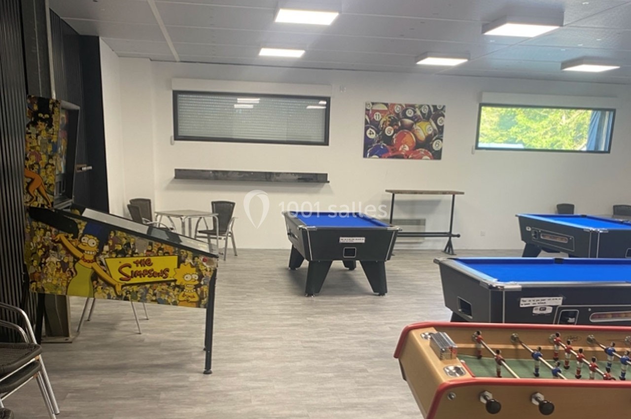 Salle de loisirs avec baby-foot, billards et flipper décoré, éclairée par des fenêtres et des luminaires au plafond.