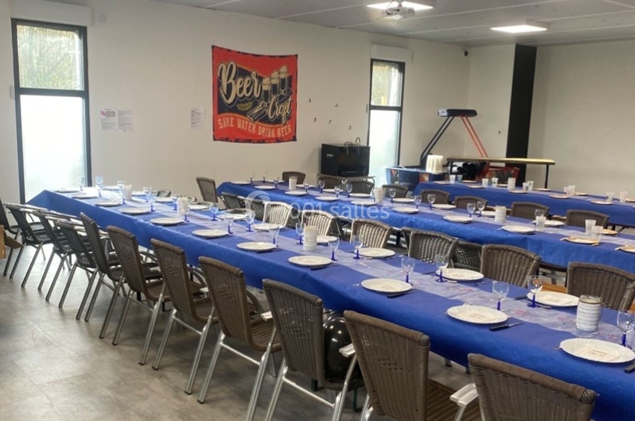 Salle aménagée avec de longues tables couvertes de nappes bleues, dressées avec assiettes et verres, prête pour un repas.