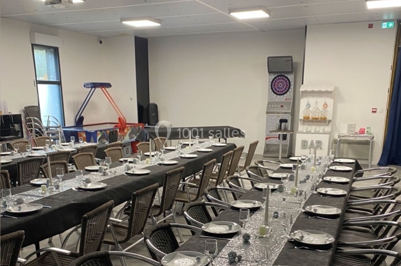Salle aménagée pour un événement avec tables dressées, chaises en osier, machine à bonbons et jeux en arrière-plan.