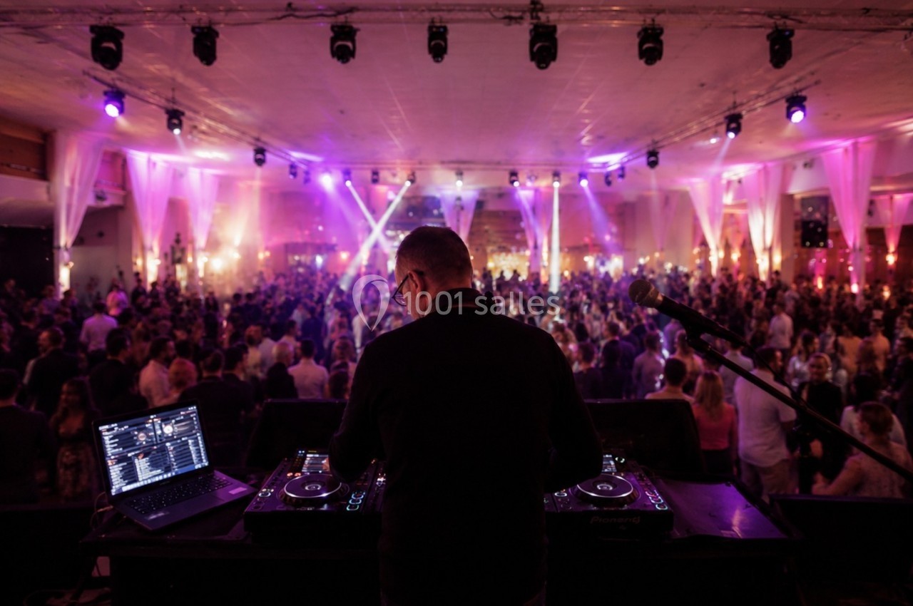 Un DJ anime une soirée dans une salle éclairée par des lumières colorées, avec une foule dansante en arrière-plan.