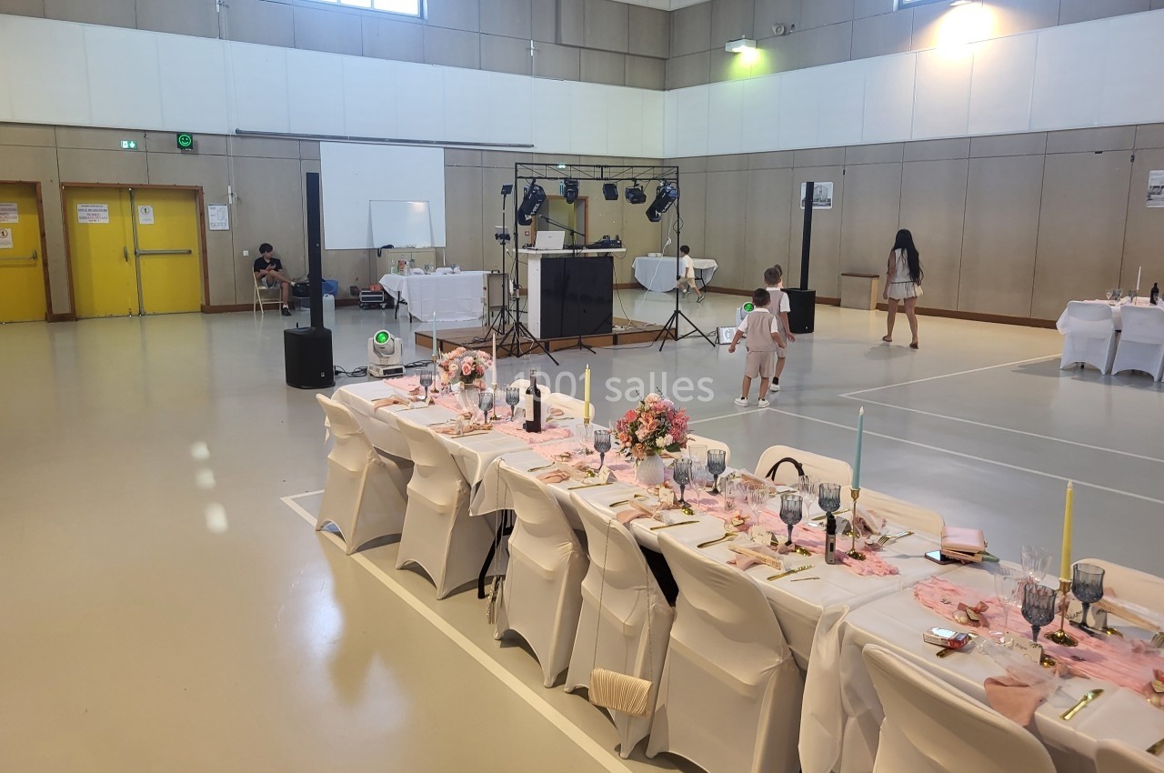 Salle de réception décorée avec une longue table dressée, des fleurs et des bougies, dans un gymnase lumineux.