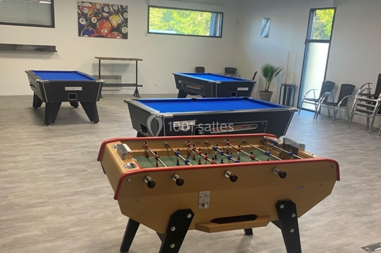Salle de loisirs avec baby-foot, billards aux tapis bleus, chaises empilées et fenêtres laissant entrer la lumière naturelle.