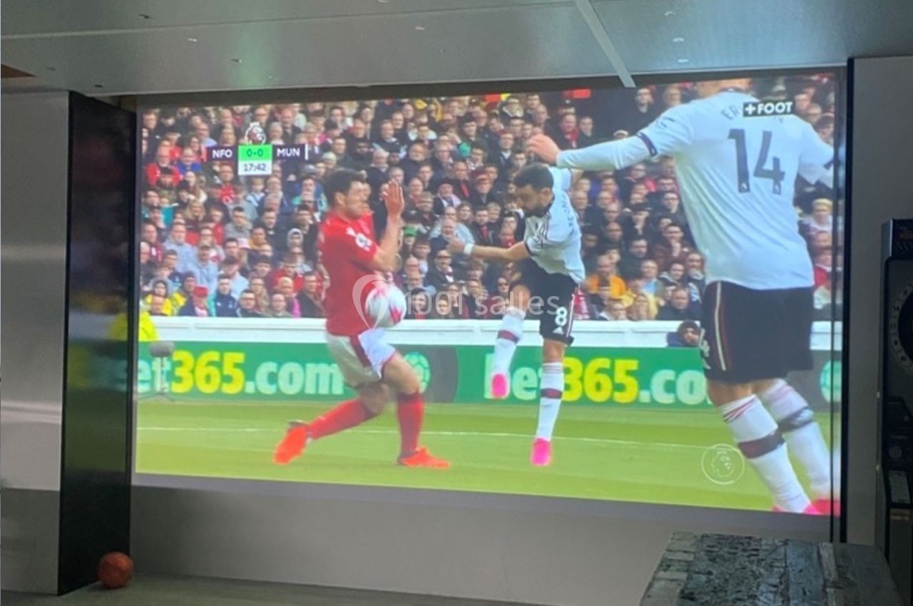 Un match de football diffusé sur un écran, montrant une action entre deux joueurs en tenue rouge et blanche.