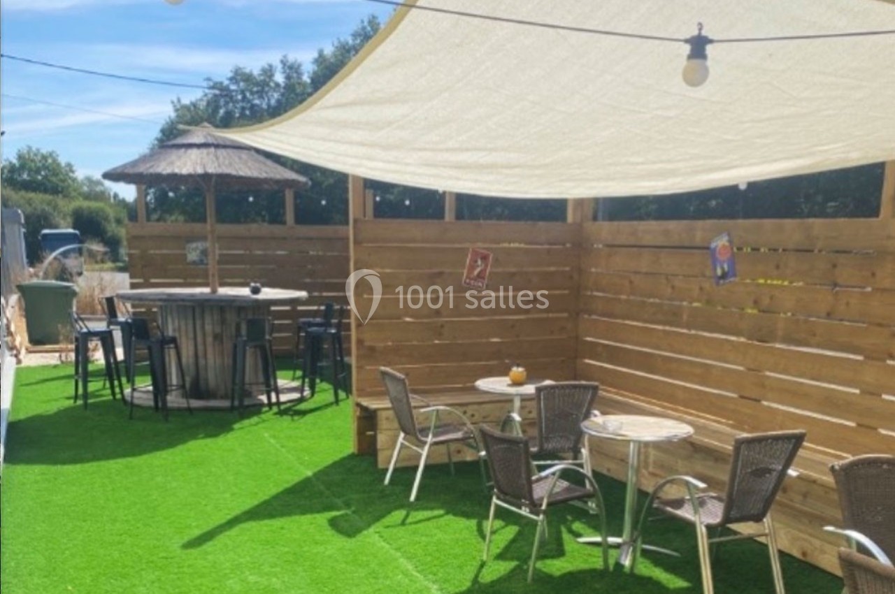Terrasse extérieure avec tables, chaises, parasol en paille et voile d'ombrage sur sol en gazon synthétique.