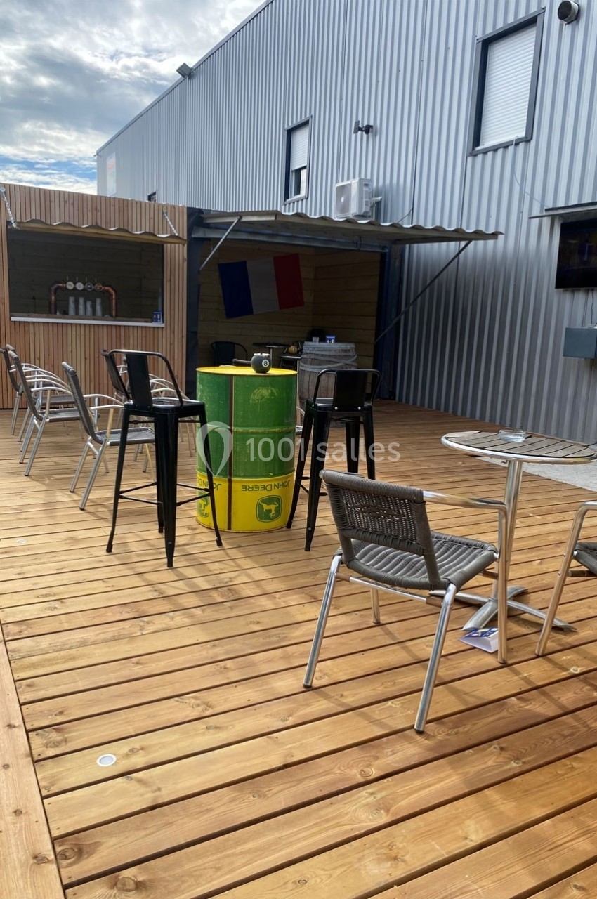 Terrasse en bois avec tables, chaises, bar en fond et tonneau vert utilisé comme table, drapeau français visible.
