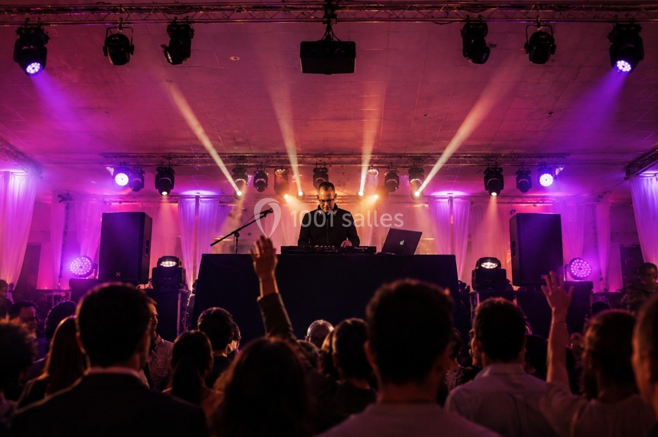 Un DJ mixe sur scène dans une salle éclairée par des projecteurs colorés, face à une foule en fête.