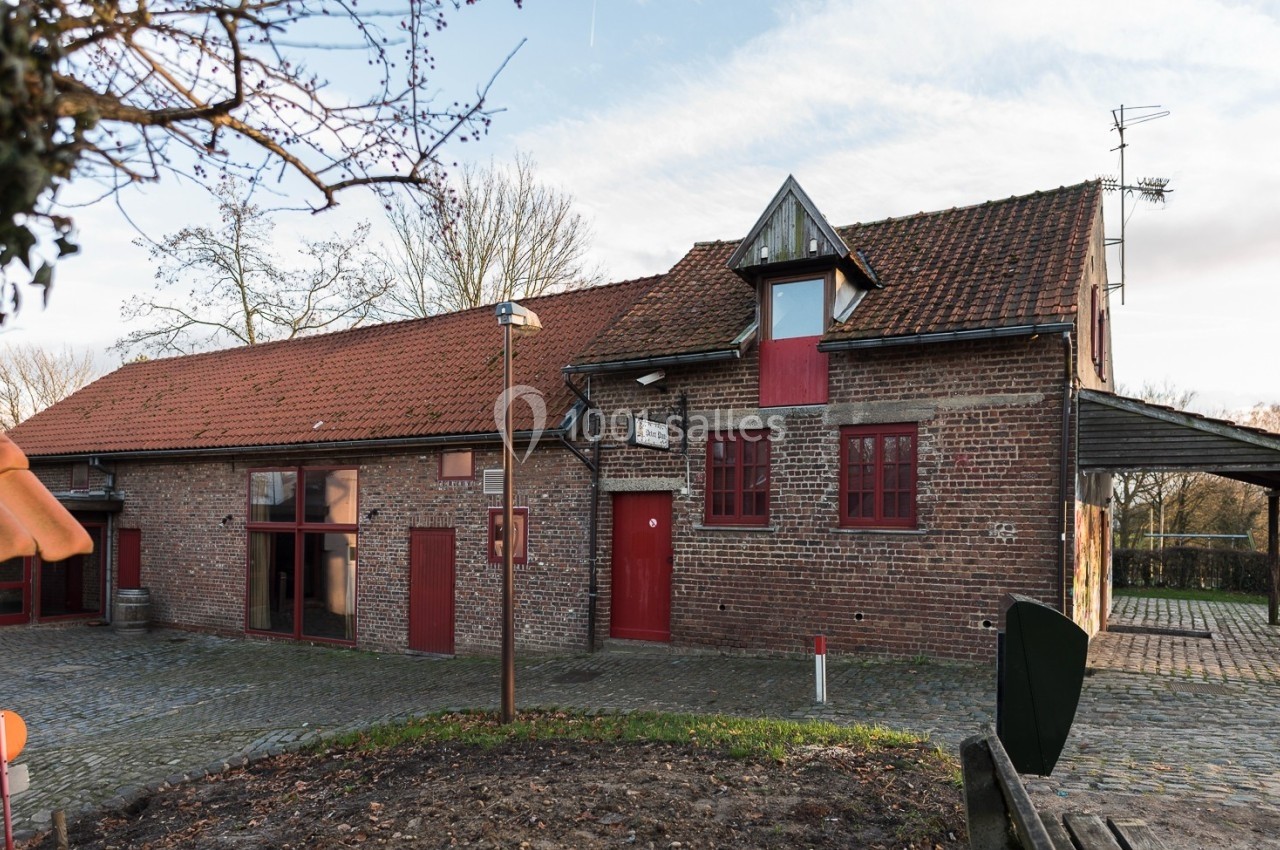 Bâtiment en briques rouges avec toit en tuiles, fenêtres rouges et petite tour, entouré de pavés et d'arbres.
