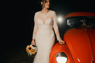 Miniature Germain Photo Reporter #6 Une femme en robe de mariée tient un bouquet de tournesols près d'une voiture ancienne rouge, de nuit.