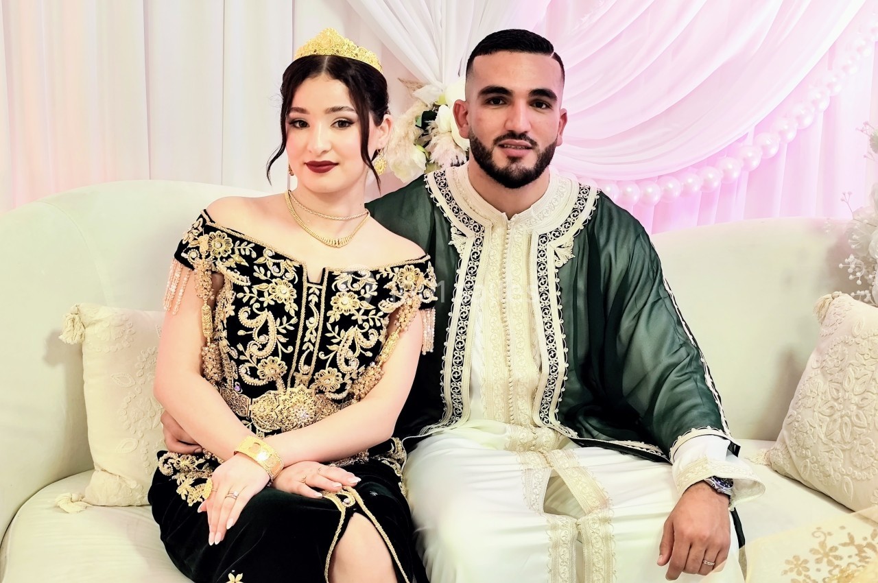 Un couple en tenue traditionnelle assis sur un canapé devant une décoration blanche et rose.