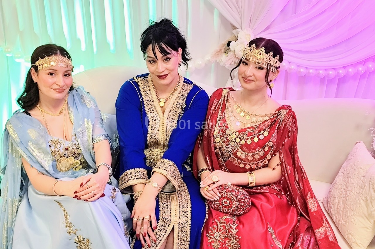 Trois femmes assises en tenue traditionnelle ornée de bijoux, posant devant un décor lumineux et drapé.