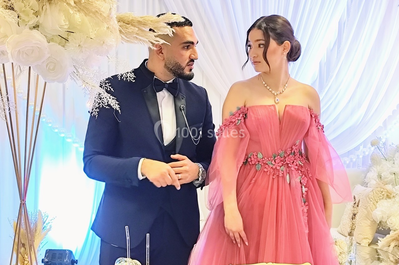 Un homme en costume et une femme en robe rose discutent devant une décoration florale blanche.