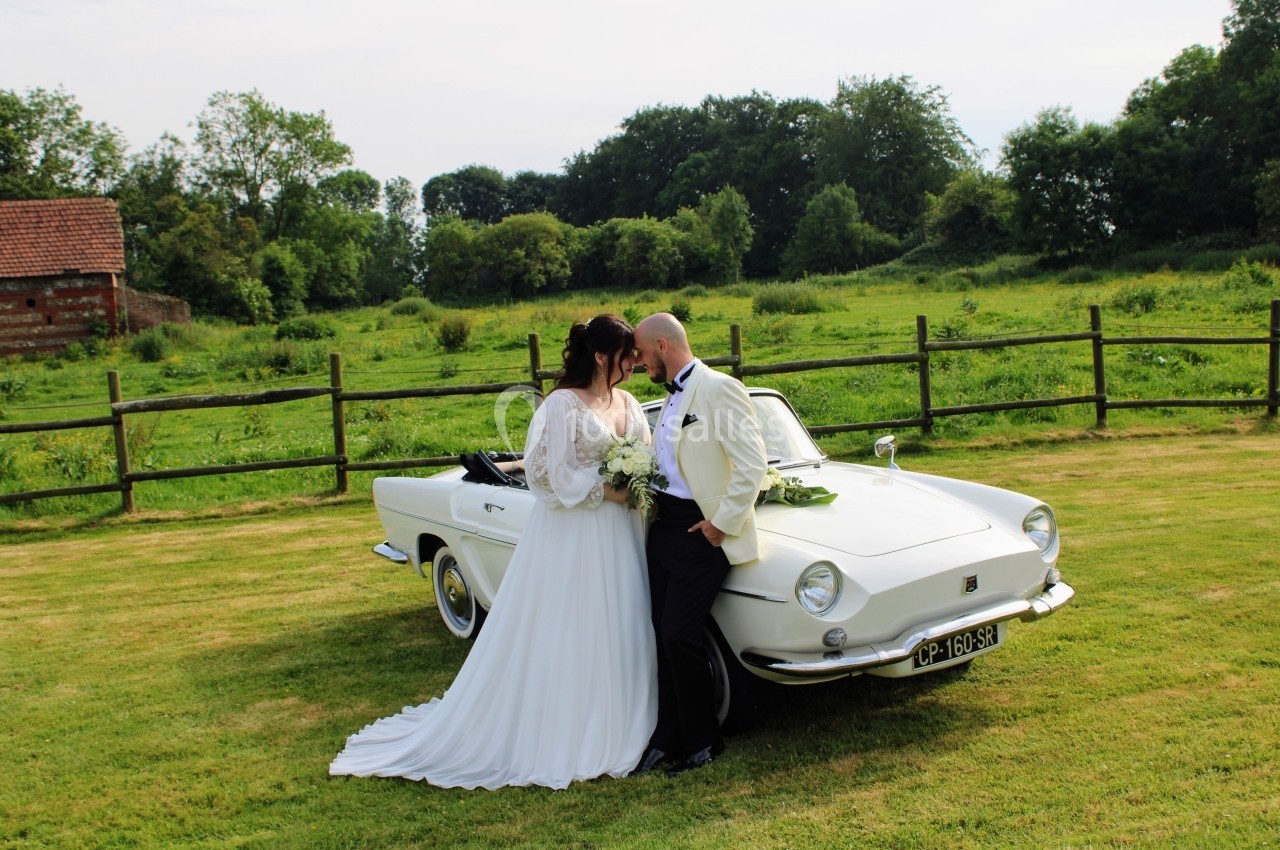 Un couple en tenue de mariage pose devant une voiture ancienne blanche dans un cadre champêtre.