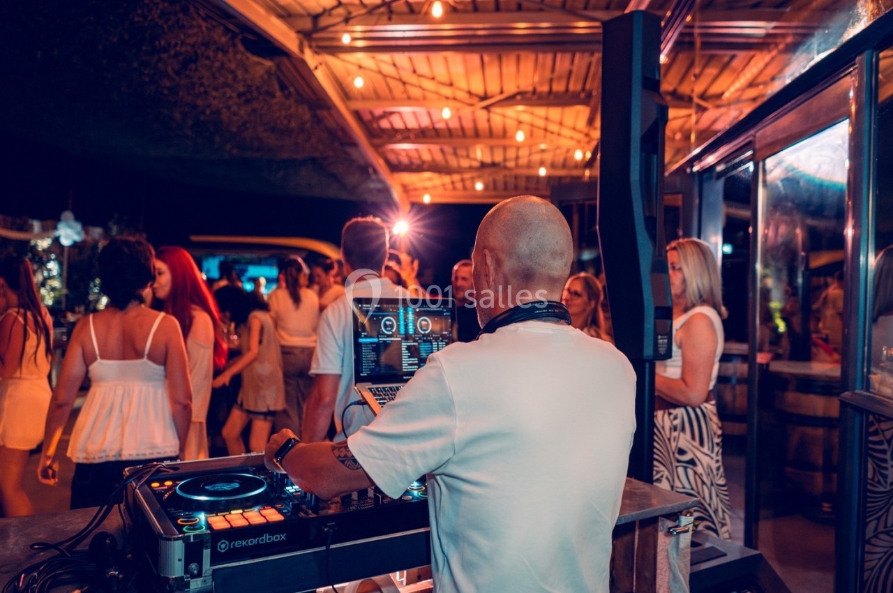 Un DJ mixe de la musique lors d'une soirée en plein air, entouré de personnes dans une ambiance lumineuse et festive.