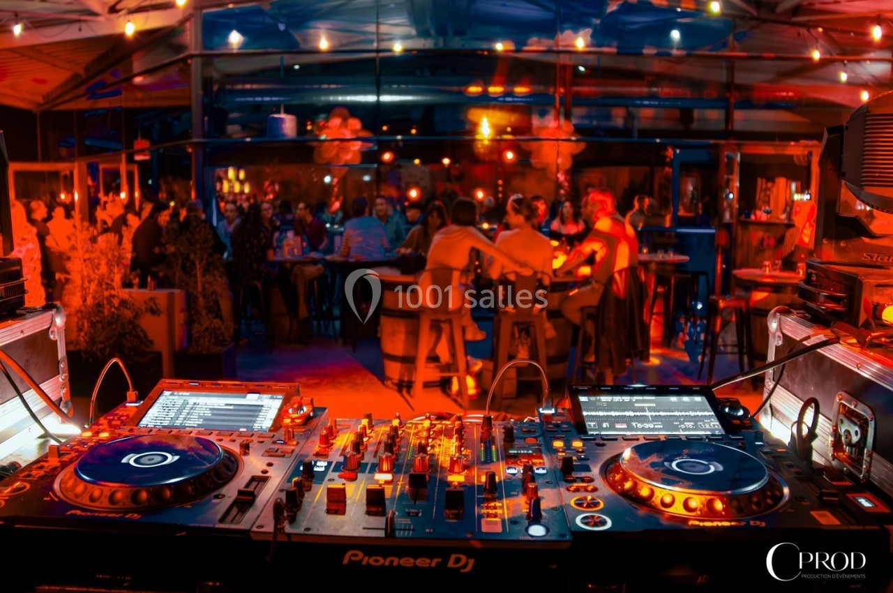 Vue d'une console de DJ en premier plan avec une ambiance festive et des personnes attablées dans un bar éclairé.