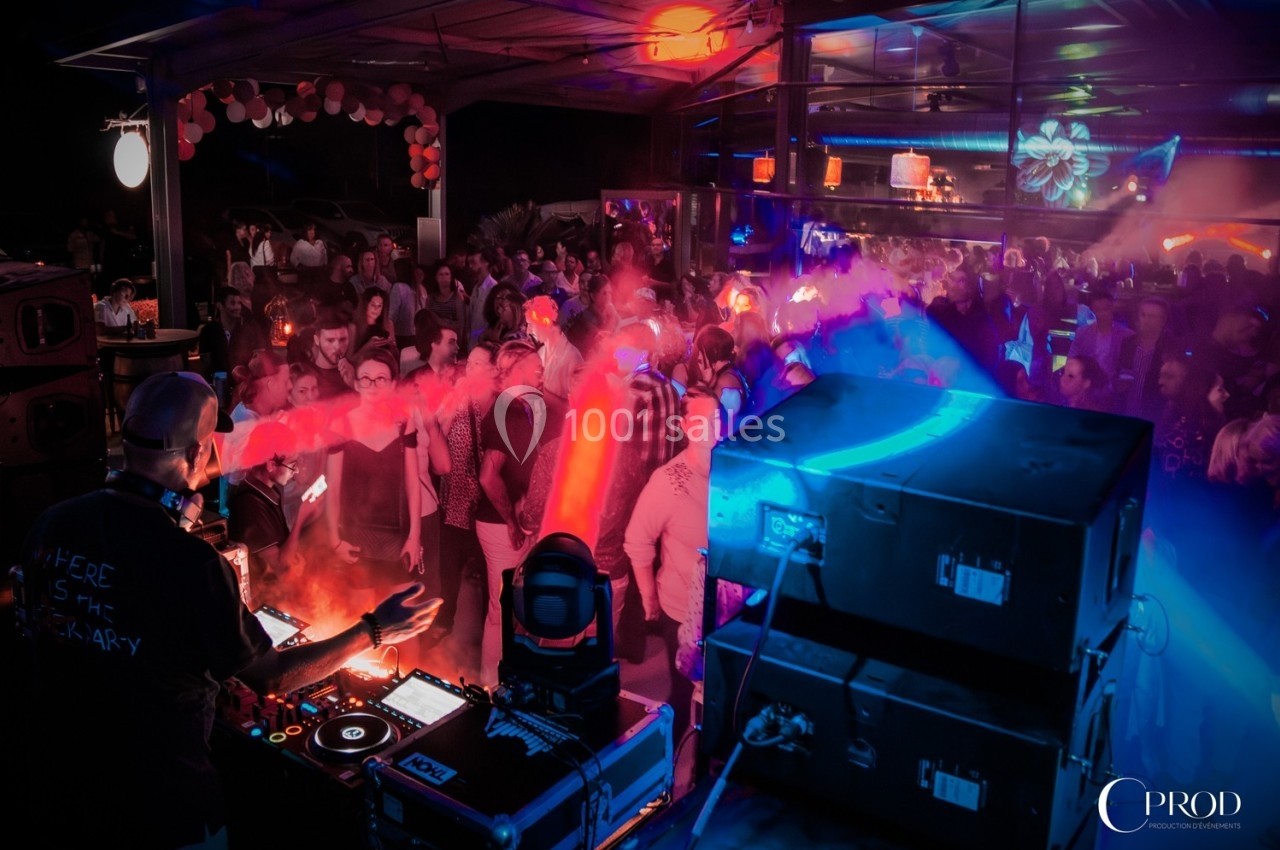 Un DJ anime une soirée dansante avec des jeux de lumière colorés devant une foule dans un espace couvert.