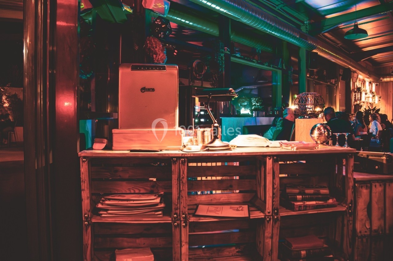 Comptoir en bois avec des étagères remplies de documents, dans un bar à l'éclairage tamisé et coloré.