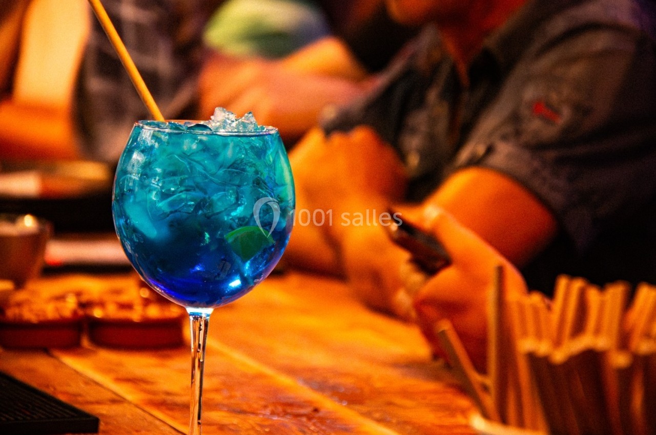 Cocktail bleu avec glaçons et une paille, posé sur une table en bois dans un bar animé.