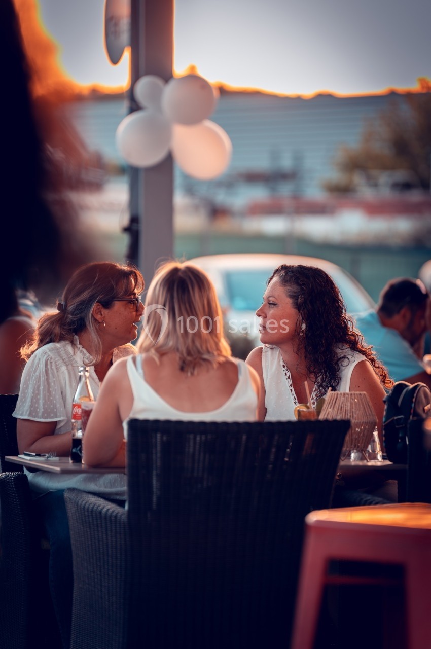 Trois femmes discutent à une table en extérieur, avec des ballons blancs et un coucher de soleil en arrière-plan.