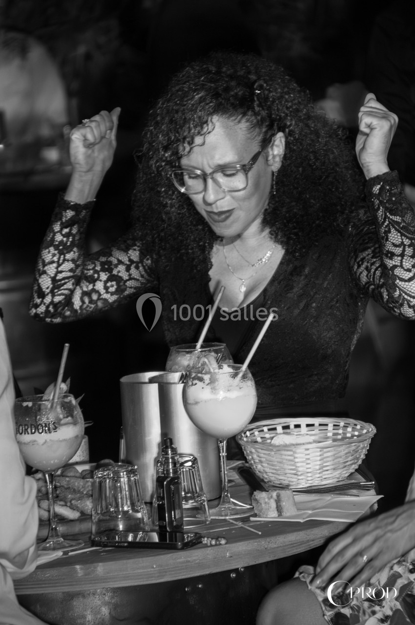 Femme portant des lunettes, levant les bras en l'air, assise à une table avec des cocktails et des paniers en osier.