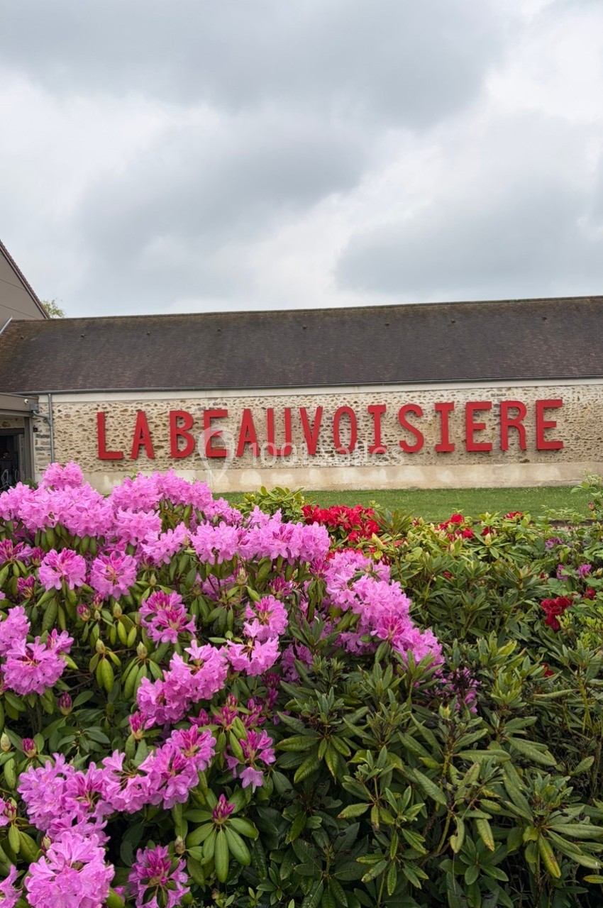 Façade en pierre avec l'inscription ’La Beauvoisière’, entourée de buissons fleuris de fleurs roses et rouges.