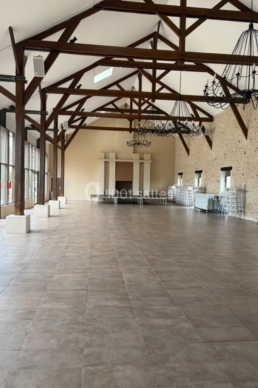 Grande salle vide avec poutres apparentes, murs en pierre, sol carrelé et grandes fenêtres laissant entrer la lumière.