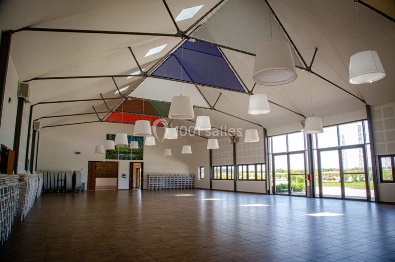Salle spacieuse avec plafond haut, luminaires suspendus, baies vitrées et chaises empilées sur les côtés.