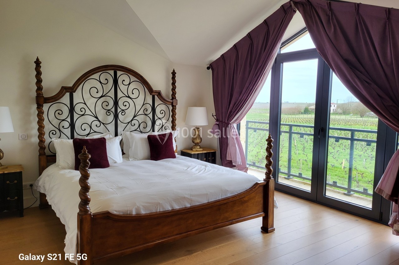 Chambre lumineuse avec lit double en bois sculpté, grandes fenêtres donnant sur un paysage de vignobles.