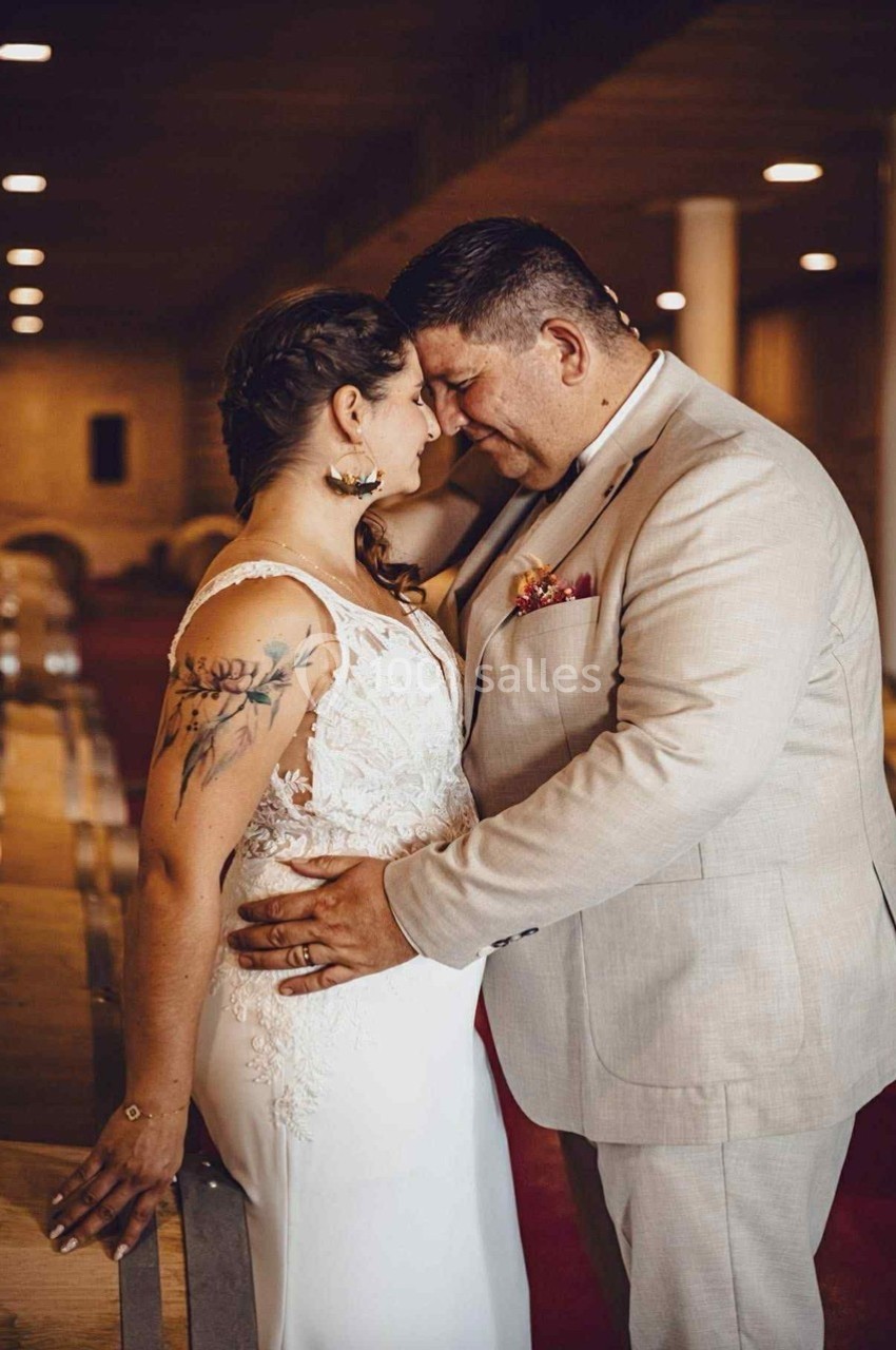 Un couple en tenue de mariage partage un moment tendre dans un lieu intérieur avec des bancs en bois.