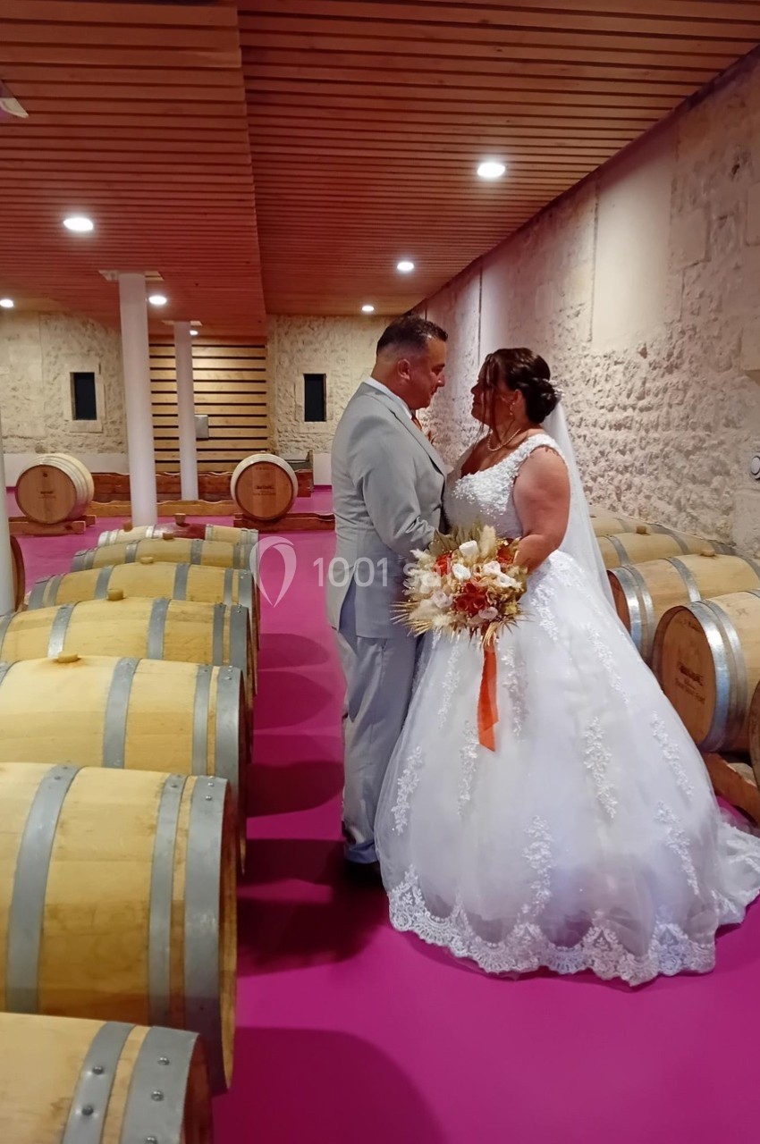 Un couple en tenue de mariage se tient entre des barriques dans une cave au sol rose.