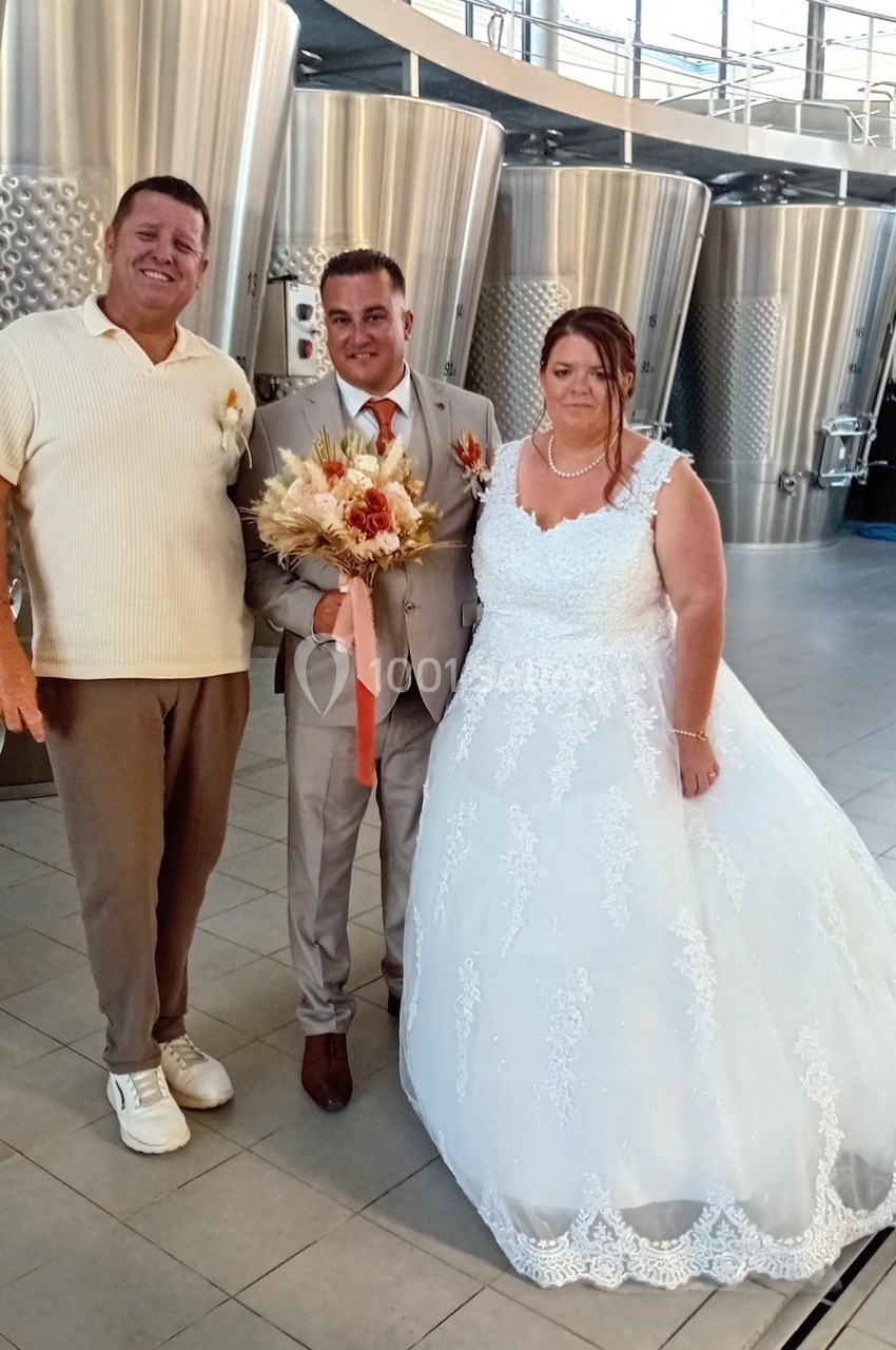 Un couple en tenue de mariage pose avec un homme dans une salle contenant de grandes cuves en acier inoxydable.