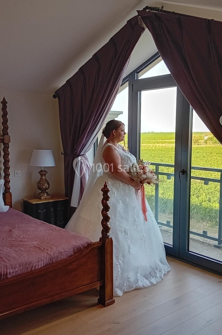 Une mariée en robe blanche tient un bouquet et regarde par une fenêtre donnant sur un paysage de vignobles.