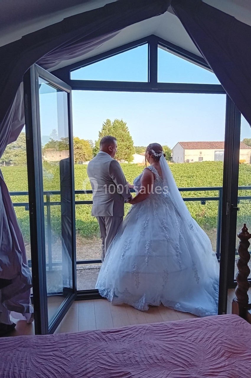 Un couple en tenue de mariage se tient sur un balcon, regardant un paysage rural avec des vignes et des bâtiments.