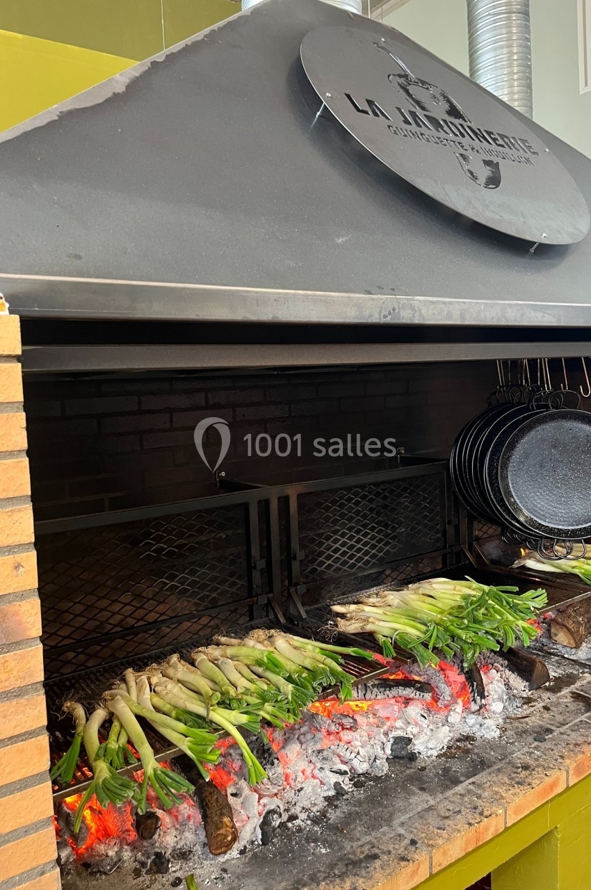 Des légumes verts grillent sur des braises rouges dans un barbecue en briques avec des poêles suspendues à côté.