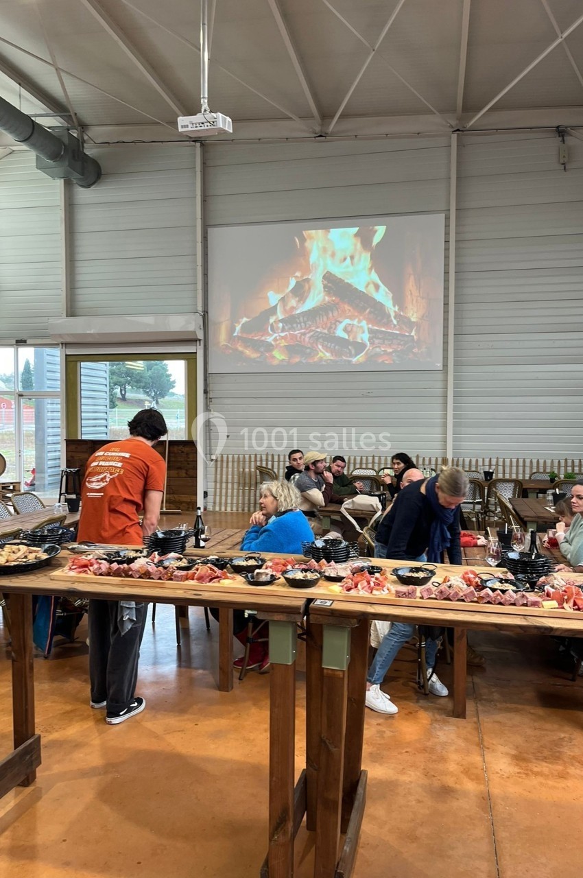 Des personnes assises autour de tables en bois garnies de plats, dans une salle avec un feu de cheminée projeté au mur.