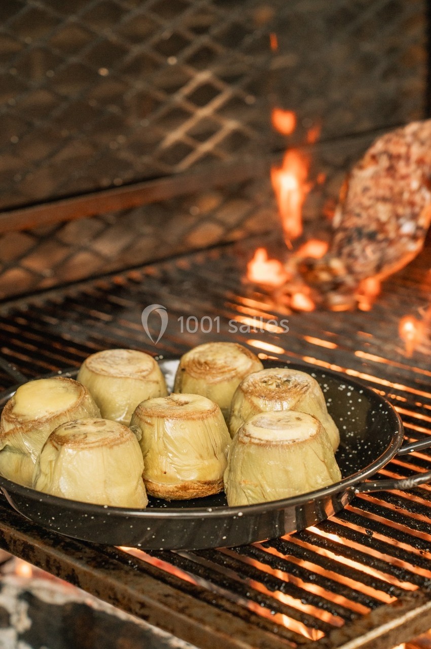 Artichauts cuits dans un plat posé sur un gril, avec des flammes et un morceau de viande en arrière-plan.