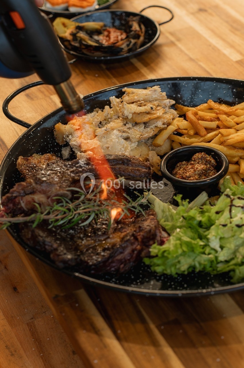 Flamme d'un chalumeau caramélisant un steak dans une assiette garnie de frites, salade et sauce.
