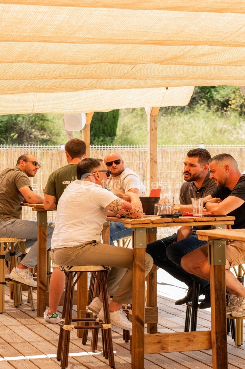 Des personnes discutent autour de tables en bois sur une terrasse ombragée par une toile tendue.