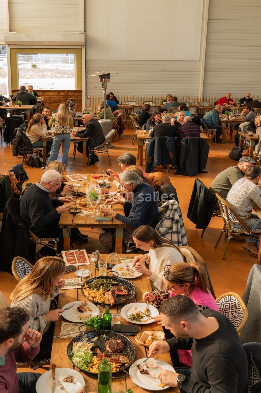 Des personnes dînent dans un grand restaurant lumineux, assises à des tables en bois, avec des plats variés partagés.