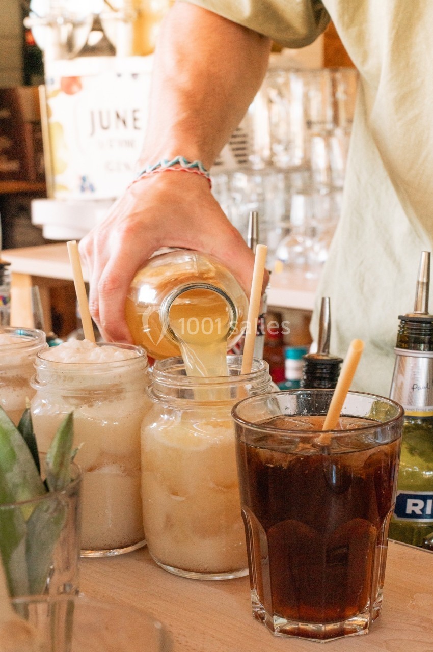 Une personne verse un liquide jaune dans un bocal rempli de glace, entouré de boissons et d'ingrédients sur un comptoir.